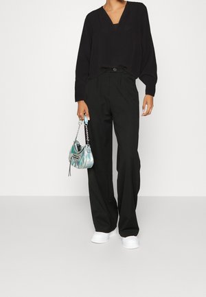 Mujer con blusa negra de manga larga y escote en V y pantalones negros de talle alto y pierna ancha, sosteniendo un bolso pequeño iridiscente, calzando zapatillas blancas.