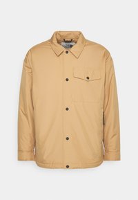 Veste couleur beige à boutons, avec rabat de poche poitrine, manches longues et col, avec le logo The North Face près de l'ourlet.