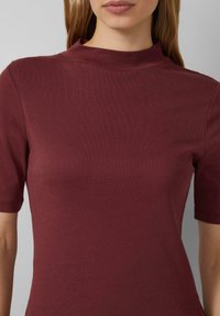 Burgundowa bluzka z krótkim rękawem i wysokim dekoltem, wykonana z gładkiego, elastycznego materiału. Charakteryzuje się dopasowanym krojem oraz brakiem widocznych wzorów lub akcentów.