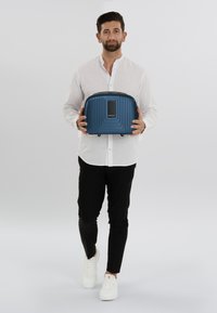 Blauwe harde cabin bagage met een verticale ribbelontwerp, handvat aan de bovenkant en merklogo. Gladde textuur met een rechthoekige vorm.