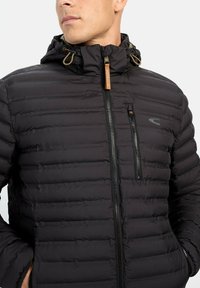 Sort pufferjakke med horisontalt quiltet design, lynlåslomme på brystet, justerbar hætte og brune læderdetaljer på lynlåshåndtagene.