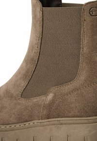 Botte Chelsea en daim marron avec panneau latéral élastique, coutures visibles et détail de semelle texturée.
