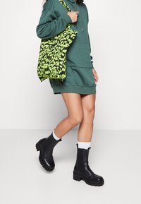 Robe sweat à capuche surdimensionnée verte, bottines noires épaisses avec semelles texturées, et un sac vert néon avec un motif noir audacieux.