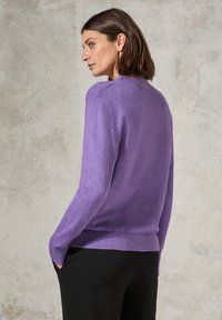 Lila stickad tröja med långa ärmar, ribbad nederkant och mudd, mjuk textur, buren med svarta byxor, sedd bakifrån.