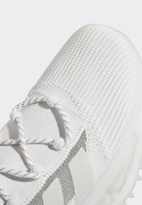 Pantofii sport albi prezintă un exterior texturat, țesut, cu șireturi rotunde, accente metalice și o talpă moale, semi-transparentă.