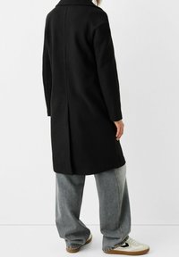 Svart ullkappa med rak silhuett, notchkrage, långa ärmar och en mitt bak-söm, kombinerad med lösa denimbyxor och sneakers.