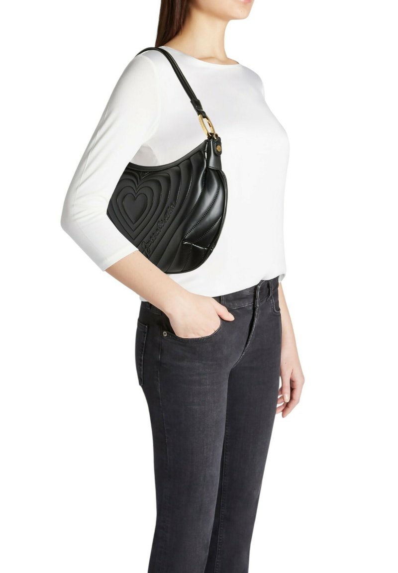 Borsa a tracolla in pelle nera con motivo a cuore trapuntato, finiture dorate e forma curva, indossata sulla spalla; abbinata a una maglietta bianca e jeans scuri.