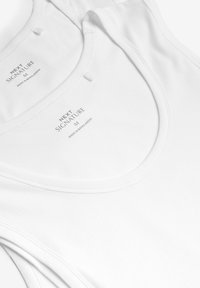 Top in cotone bianco senza maniche con scollatura rotonda, morbido al tatto e etichette di marca visibili che riportano "NEXT SIGNATURE M" e "MADE IN BANGLADESH."