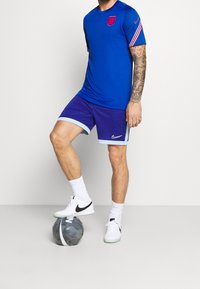 Blå Nike-t-shirt med röd krage och randiga ärmar, tillsammans med lila shorts med ljusblåa detaljer. Vita sneakers; boll vid fötterna.