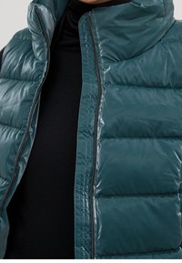 Veste matelassée bleu sarcelle avec un col montant, une texture brillante et une fermeture éclair, portée par-dessus un haut noir. Caractérisée par des coutures matelassées pour l'isolation.