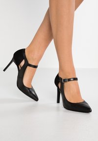 Černé vysoké podpatky s špičatou špičkou, výřezovými vzory a tenkým zápěstním páskem se malou přezkou. Vyrobeno z hladké kůže.