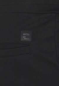 Svart tyg med en liten rektangulär Alpha Industries-logotyp på en patch ovanför en horisontell ficka.