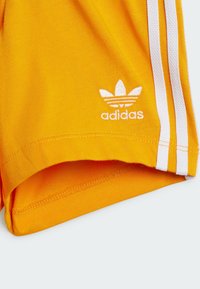 Shorts de sport jaunes en tissu lisse, avec des bandes blanches sur les côtés et un petit logo Adidas blanc brodé près de l'ourlet.
