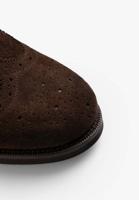 Zapato de ante marrón con perforaciones decorativas, punta redondeada y un ribete cosido a lo largo de la suela. La superficie texturizada añade profundidad.