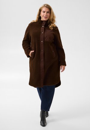 Femme aux longs cheveux blonds porte un long manteau marron foncé avec des boutons-pression, un pantalon bleu et des bottines noires, debout avec une main dans la poche.