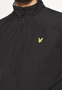 Zwarte ritsjack gemaakt van lichtgewicht materiaal. Heeft een staande kraag en een gele geborduurde logo op de borst. Soepele textuur.