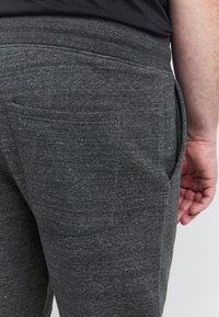 Sötétszürke, texturált anyagból készült melegítőnadrág, hátulján zsebbel és rugalmas derékrésszel, kézzel látható hátulnézetből.