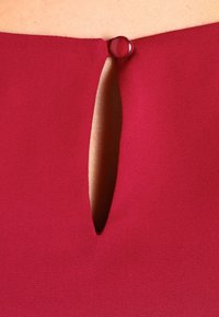 Détail d'une robe bordeaux avec une ouverture en forme de goutte d'eau au dos, fermée par un bouton rond rouge. Texture lisse avec une finition douce.