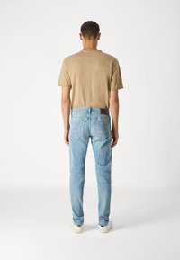 Hombre que lleva vaqueros de un azul claro, camiseta beige y zapatillas blancas, de pie y de espaldas en un fondo blanco liso.