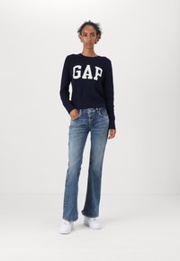 Maritimer Strickpullover in Navy mit einem weißen "GAP" Logo, kombiniert mit hellblauen Schlagjeans und weißen Sneakers. Einfaches, lässiges Design, strukturierte Passform.
