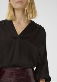 Zwarte blouse met een V-hals, plooi-detail in het midden en lange, losse mouwen. Het materiaal heeft een gladde, licht glanzende textuur.