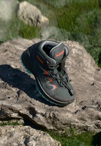 Grau Wanderschuh mit lebhaften blauen und orangefarbenen Akzenten, ausgestattet mit einem gepolsterten Kragen, strukturiertem Obermaterial und rutschfester Sohle, die auf einem Felsen platziert ist.