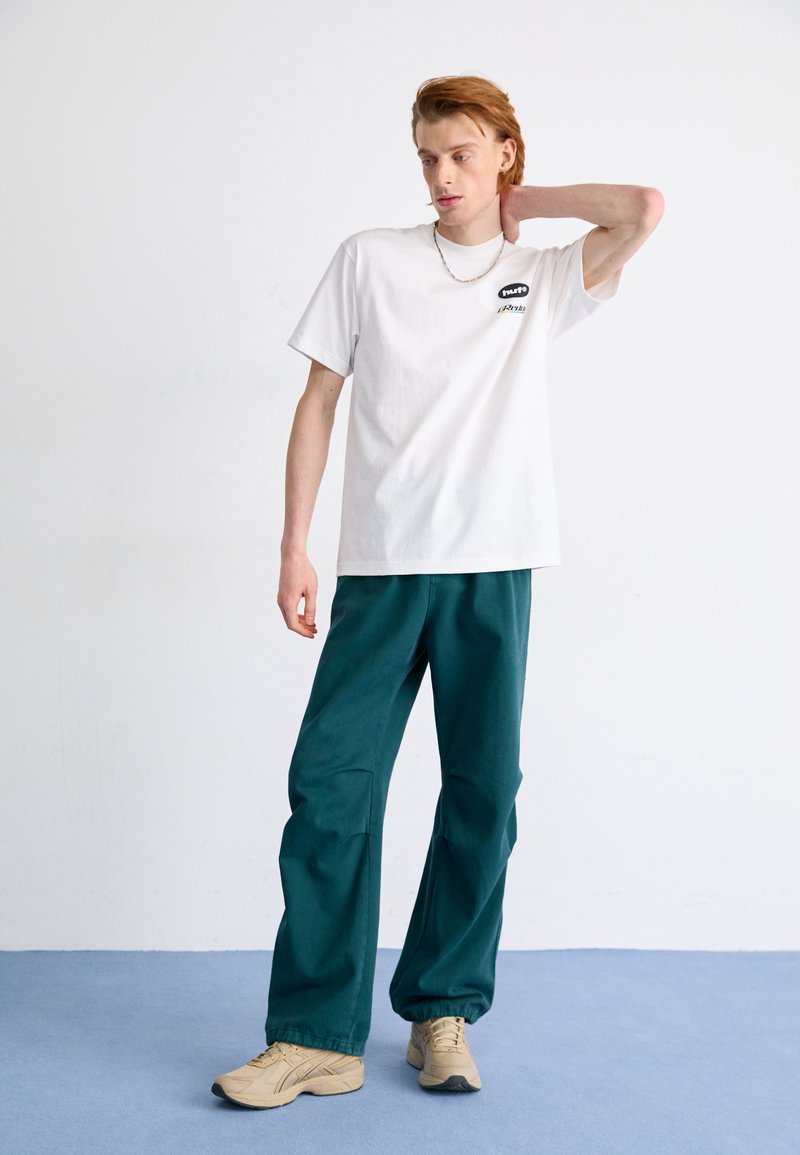 T-shirt en coton blanc avec logo noir, associé à un pantalon teal à coupe décontractée. Des baskets beiges complètent la tenue. Lignes épurées et design décontracté.