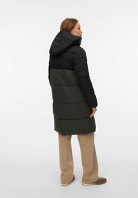 Paminkštintas paltas, kurio viršutinė dalis juoda, o apatinė tamsiai žalia, su kapišonu ir vertikaliomis siūlių detalėmis. Derinamas su smėlio spalvos kelnėmis.