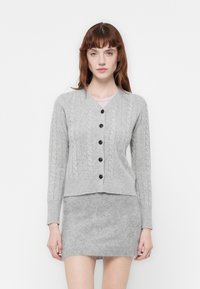 Cardigan grigio lavorato a maglia con un motivo a trecce, caratterizzato da un collo a V, maniche lunghe e bottoni scuri. Indossato sopra una gonna grigia abbinata.