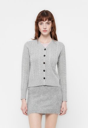 Cardigan tricoté gris avec un motif en cable, présentant un col en V, des manches longues et des boutons foncés. Porté sur une jupe grise assortie.