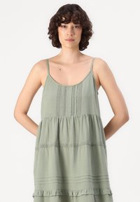 Φωτεινό πράσινο αμάνικο φόρεμα με ρυθμιζόμενους straps, που διαθέτει υφές σε επίπεδα και δαντελένιες λεπτομέρειες κατά μήκος του κορμού και του στριφώματος.