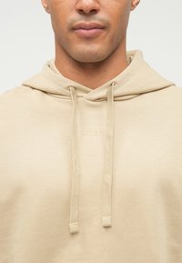 Primo piano di una persona che indossa un hoodie beige con stringhe e un piccolo logo "HUGO" sul petto.