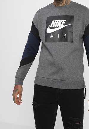 Personne portant un sweat gris Nike Air avec des manches noires et bleu marine et un jean noir déchiré, visible du cou aux cuisses.