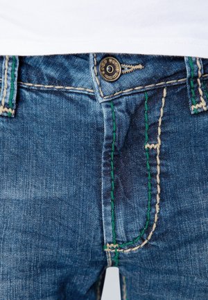 Forfra visning af blå denimjeans med kontrasterende grønne og beige syninger, en frontlomme og en metallukning med mærke.