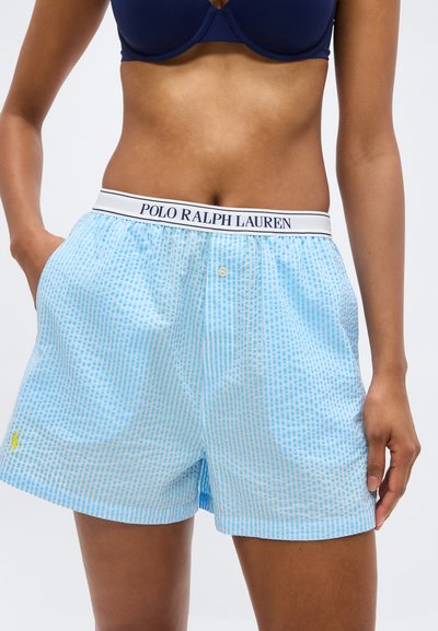 Γυναίκα που φοράει ανοιχτό μπλε και λευκά ριγέ σορτς boxer Polo Ralph Lauren και σκούρο μπλε σουτιέν, στέκεται με τα χέρια στις τσέπες των σορτς.