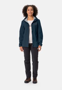 Giacca impermeabile blu marina con cappuccio grigio, chiusura lampo frontale completa e due tasche laterali, abbinata a pantaloni neri e stivali da trekking marroni.