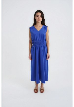 Frau mit langen dunklen Haaren in einem ärmellosen blauen Midikleid und braunen Sandalen steht vor einem einfarbigen hellen Hintergrund.