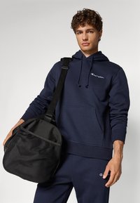 Marineblauer Hoodie mit Kängurutasche und Kordelzügen; schwarze Tasche mit Netzpanel; beide zeigen das Champion-Logo in Weiß.
