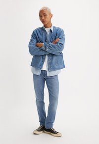 Joven con cabello corto rubio se encuentra de brazos cruzados, vistiendo una chaqueta de denim en capas, jeans, camiseta blanca y zapatillas negras, frente a un fondo blanco.