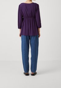 Lila blus med trekvartslånga ärmar och gathered midja, matchad med löst sittande blå denimbyxor. Svarta platta skor är på. 