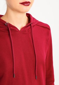 Sudadera granate con capucha ajustable, agujas metálicas y textura suave. Presenta un diseño simple con mangas raglán y un corte relajado.