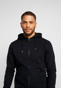 Sort lynlås hoodie med snorhætte, kengurulomme og glat, blød tekstur. Enkelt design med sølv lynlåsdetaljer.