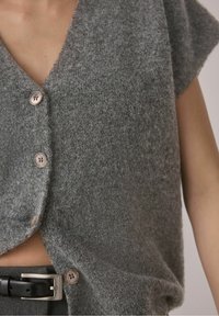 Gros plan d'une personne portant un gilet en laine gris boutonné sur un pantalon gris avec une ceinture noire, montrant la texture et les boutons.