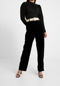 Frau trägt schwarzes, durchscheinendes, gestreiftes Langarm-Top, schwarze Samthose mit hoher Taille, beigen Gürtel mit runder Schnalle und schwarze Absatzschuhe mit offenen Zehen.