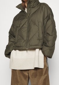 Přikrývková olivově zelená péřová bunda s vysokým límcem, zapínáním na zip a diagonálním prošíváním. Nošená přes světlý žebrovaný svetr a hnědé manšestrové kalhoty.