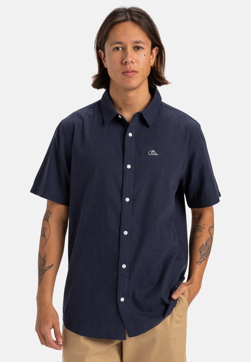 Camisa de botones de manga corta en color navy, confeccionada en tela tejida, con un pequeño logo blanco en el pecho y botones blancos en la parte delantera.