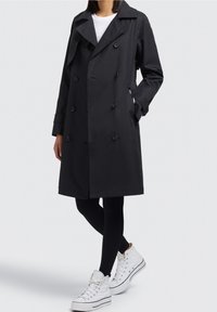 Trench-coat noir à double boutonnage avec de larges revers, poches latérales et coupe droite. Porté avec des leggings noirs et des baskets blanches montantes.