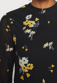 Blusa negra con mangas largas transparentes, que presenta un patrón floral en blanco y amarillo con acentos de azul y pequeñas formas triangulares.
