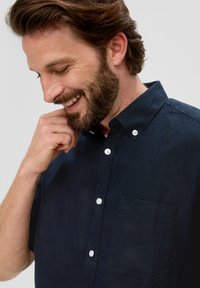 Navyblauwe, kortekogelige button-up shirt gemaakt van gestructureerde stof, met een patchzak en witte knopen langs de sluiting.