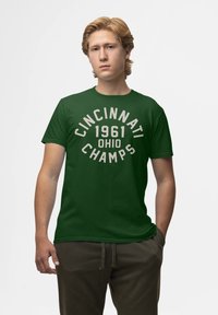Joven con cabello rubio que lleva una camiseta verde con el texto blanco "Cincinnati 1961 Ohio Champs", de pie contra un fondo claro y liso.